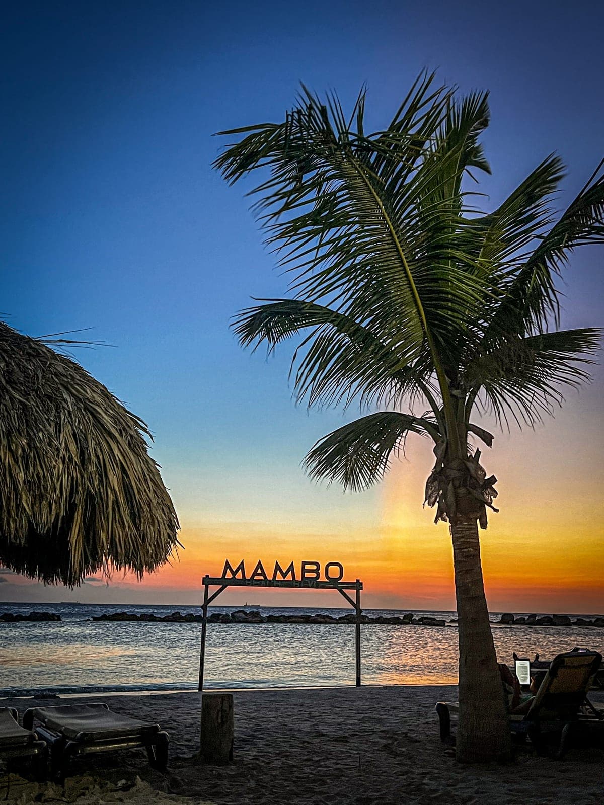 Zonsondergang bij het Mambo Beach-bord met palmboom en rieten parasol op het strand